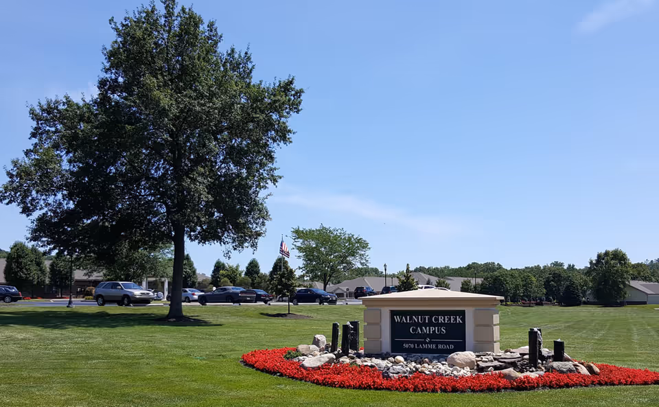 Entrance sign for Walnut Creek Campus on a landscaped lawn with a large tree and campus buildings in the background.