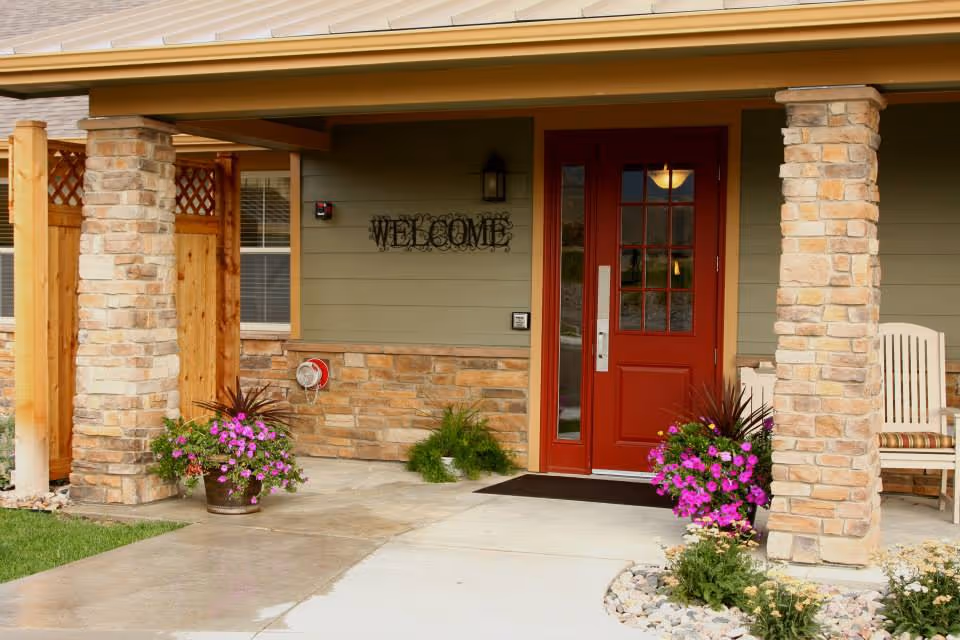 Front entrance of a senior living facility with a red door, stone columns, and flowers.