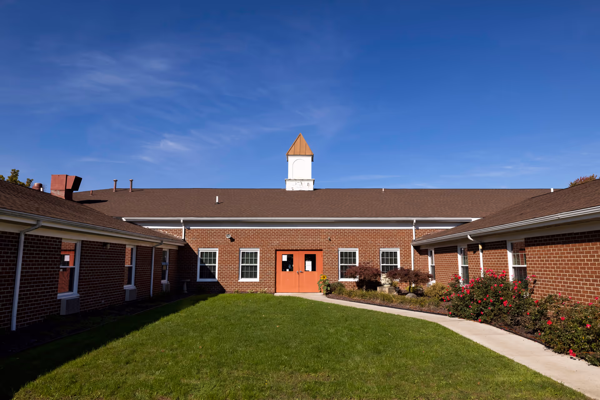Courtyard view of a single-story brick senior care building with a central orange door, cupola, lawn and walkway.