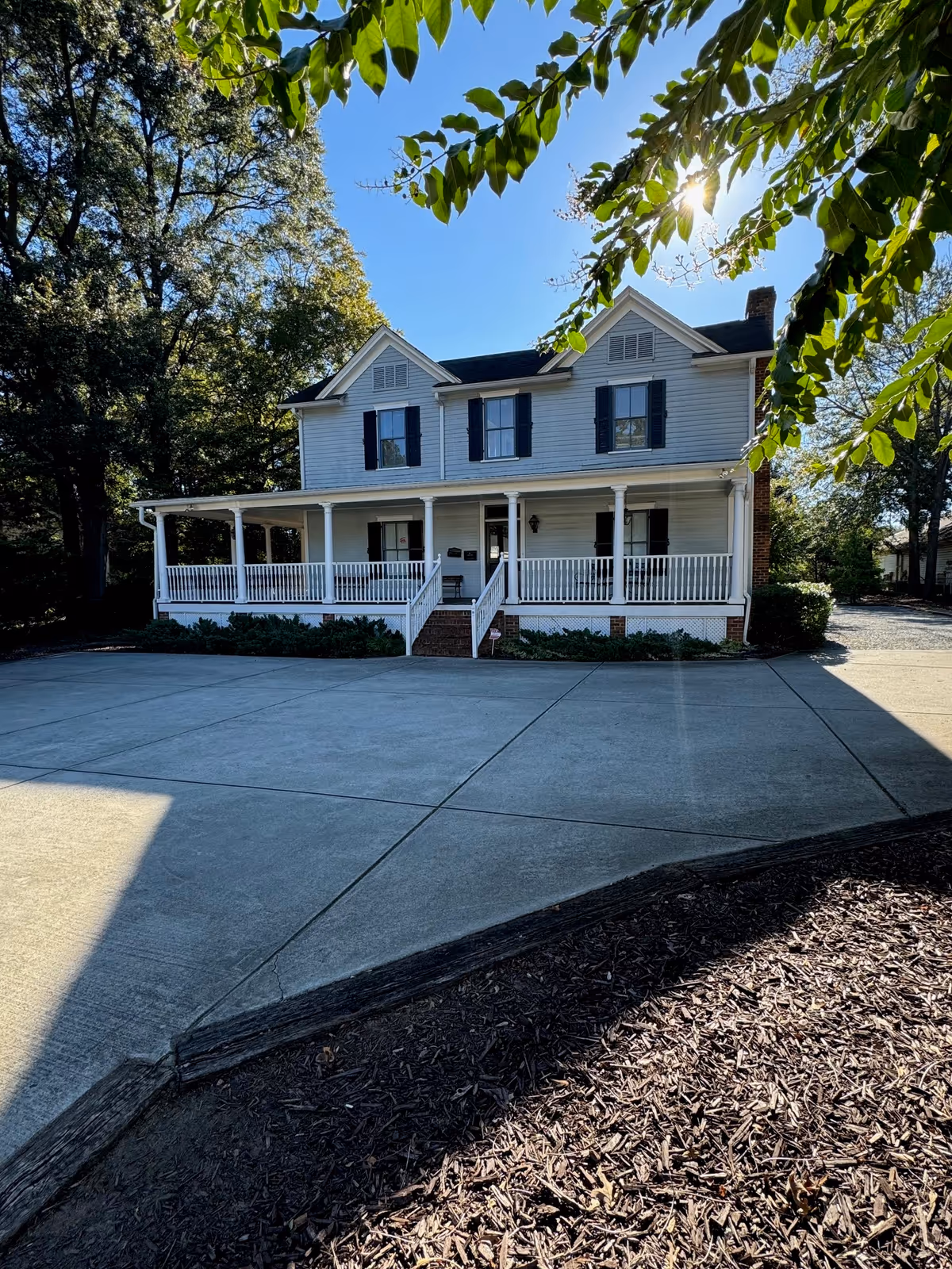 White two-story house with a wraparound porch and black shutters on a paved driveway.