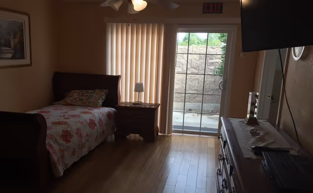 A bedroom with a single bed, floral quilt, nightstand, dresser, and sliding glass door.