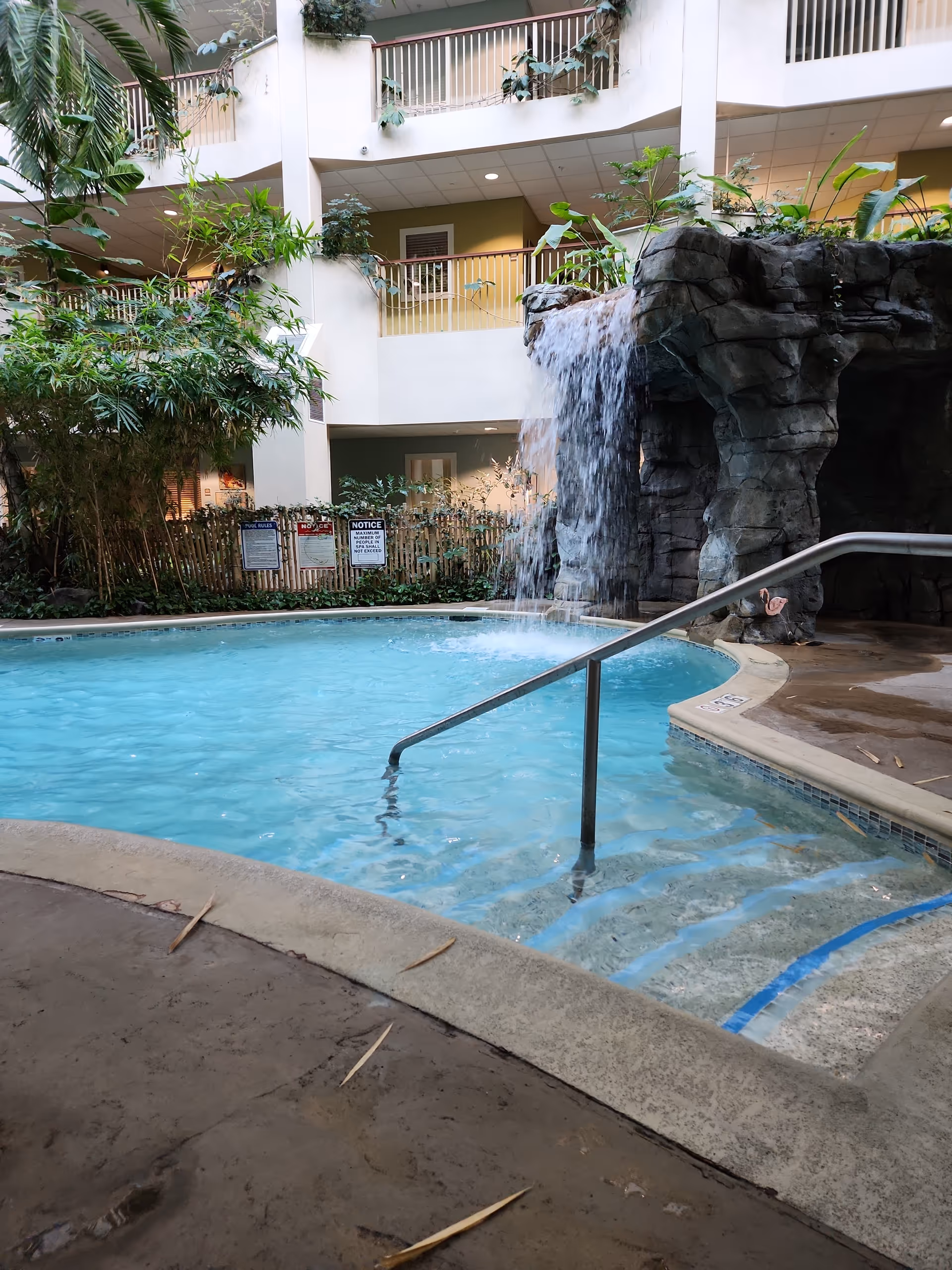 Indoor atrium pool with waterfall feature, lush plants, and multi-level balconies surrounding the area.