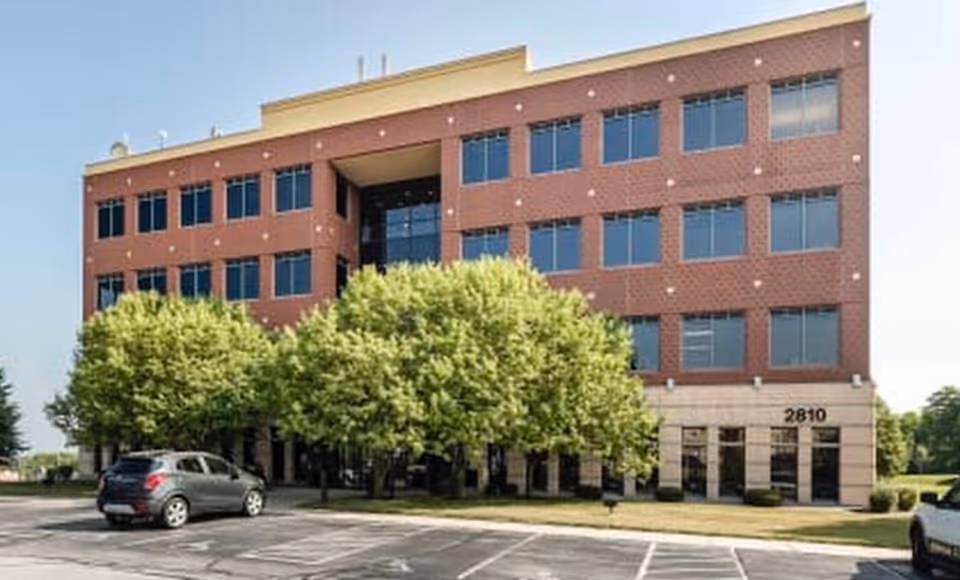 Three-story brick building with large windows, trees, and a parking lot in front.
