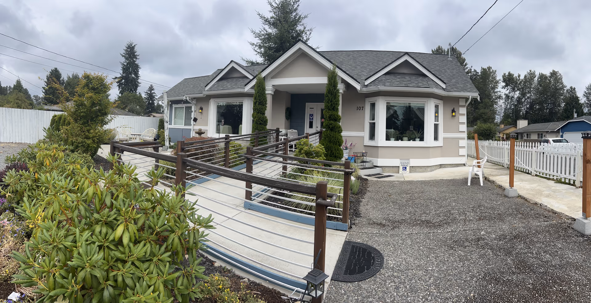 Front exterior view of a single-story senior care home with ramp, landscaping, and white fences.