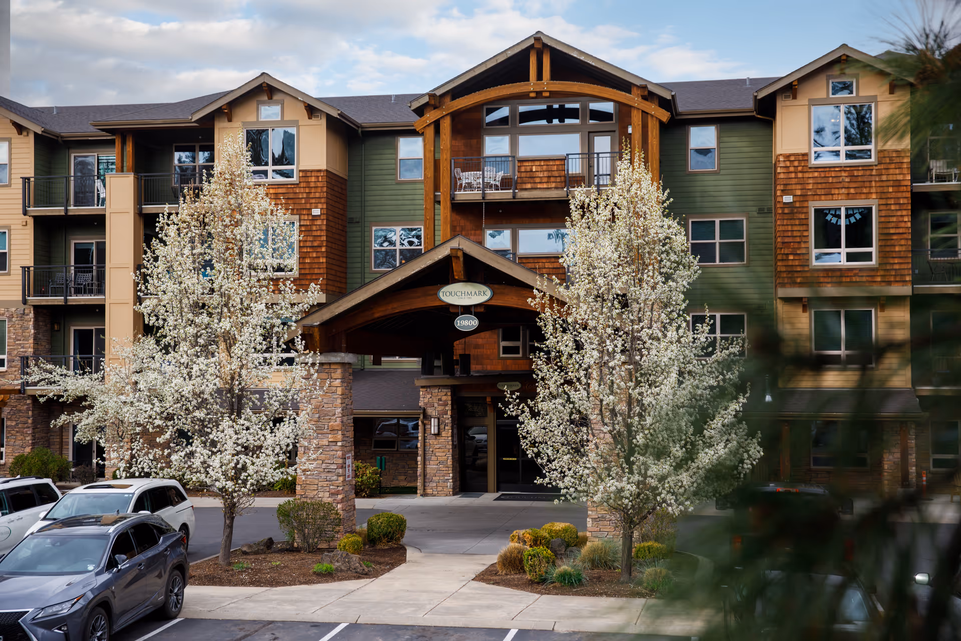 Front exterior view of Touchmark at Mount Bachelor Village, a multi-story building with green and brown siding, stone pillars, and a covered entrance. Two blossoming trees flank the entrance, and several cars are parked in front.