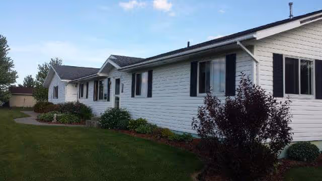 Exterior view of a single-story white-siding building with shrubs, a manicured lawn, and a clear blue sky.