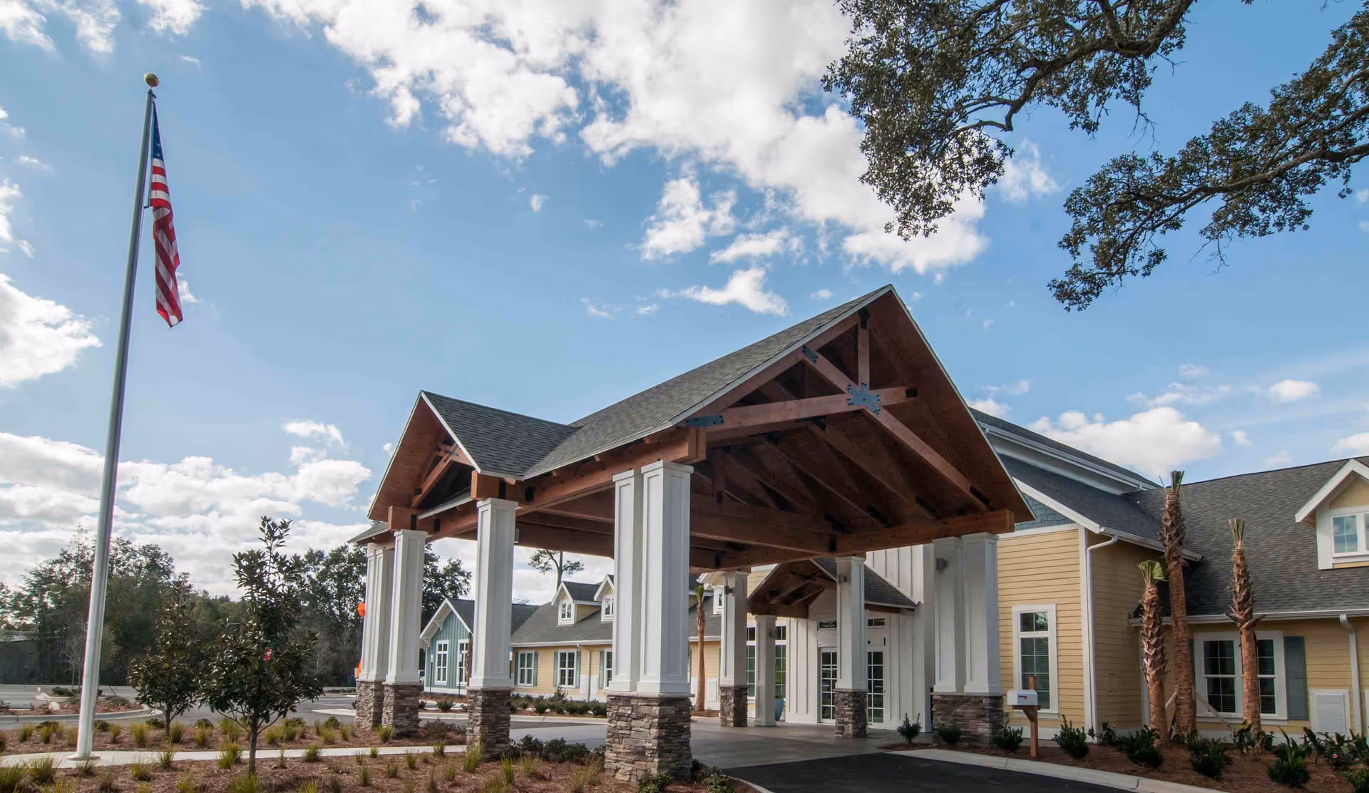Exterior view of a senior living facility with a covered entrance supported by white columns with stone bases, an American flag on a flagpole, and a partly cloudy sky.