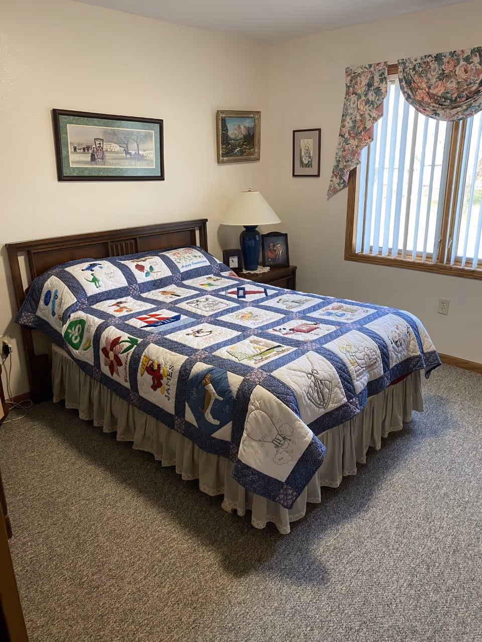 A cozy bedroom with a bed covered by a colorful patchwork quilt featuring various designs. The room has beige walls with three framed pictures hanging above the bed. A wooden nightstand with a blue lamp and a framed photo sits next to the bed. A window with vertical blinds and floral valance lets in natural light. The floor is carpeted in a neutral tone.