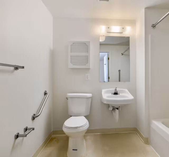 Small apartment bathroom with toilet, sink, mirror, and safety grab bars.
