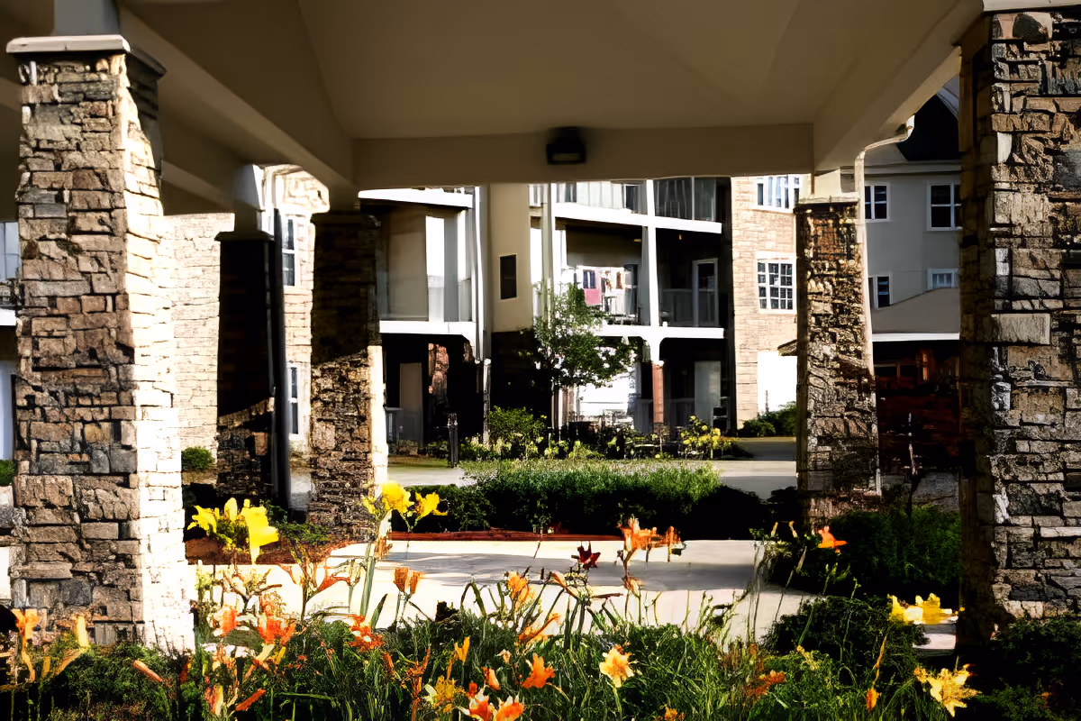 Covered stone-pillared entrance and landscaped flower beds in front of a senior living building and courtyard.