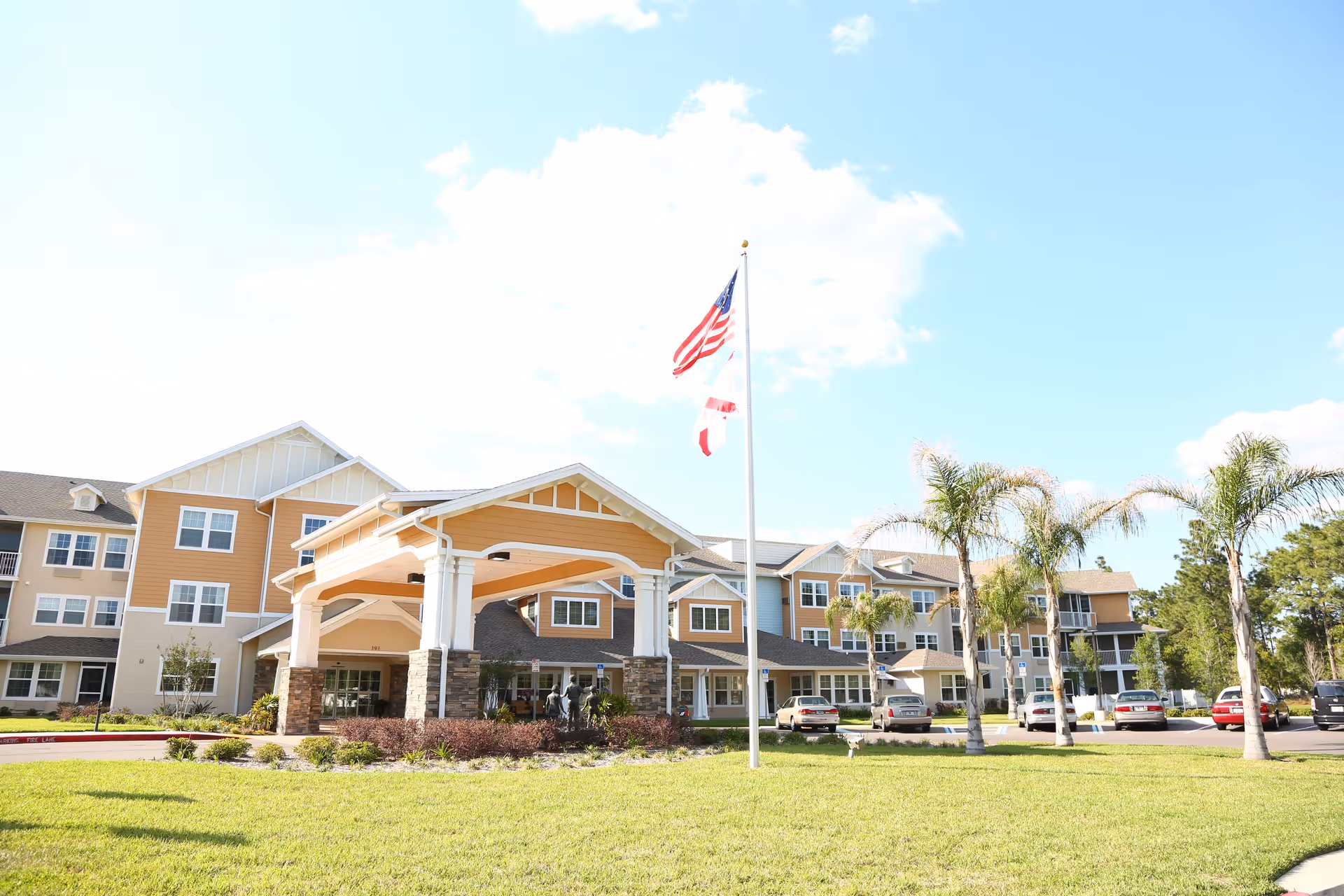 Exterior view of a multi-story senior living facility with a covered entrance, several parked cars, palm trees, and an American flag flying on a flagpole in front of the building under a partly cloudy sky.