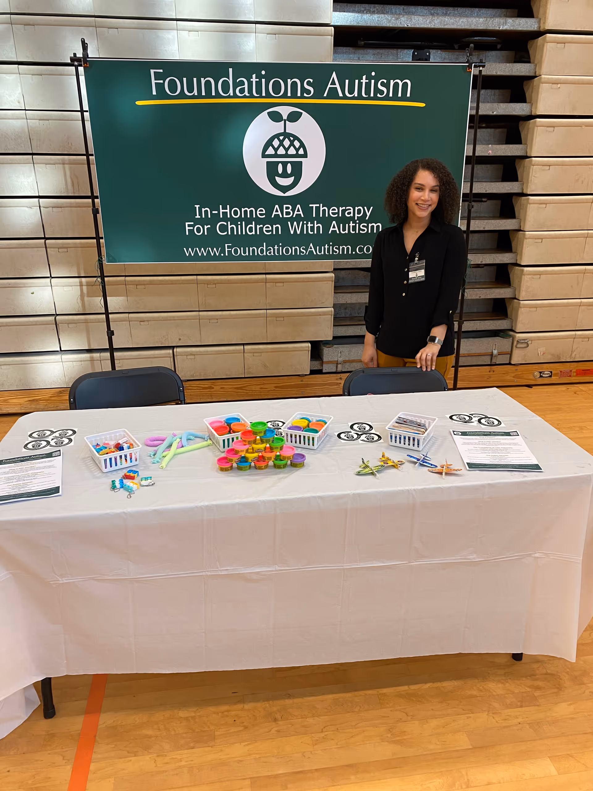 A staff member stands beside a Foundations Autism banner at a toy display table indoors.