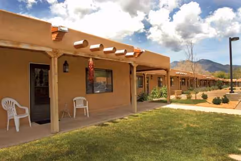 Adobe-style, single-story facility exterior with a shaded porch, white chairs, and a grassy lawn.