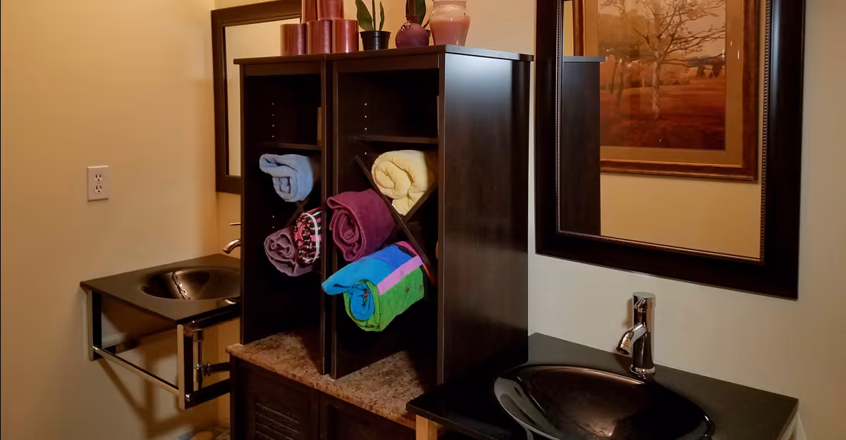 Interior view of a bathroom featuring two black sinks with chrome faucets, a dark wooden cabinet with rolled colorful towels, a large mirror, and a framed picture on the wall.