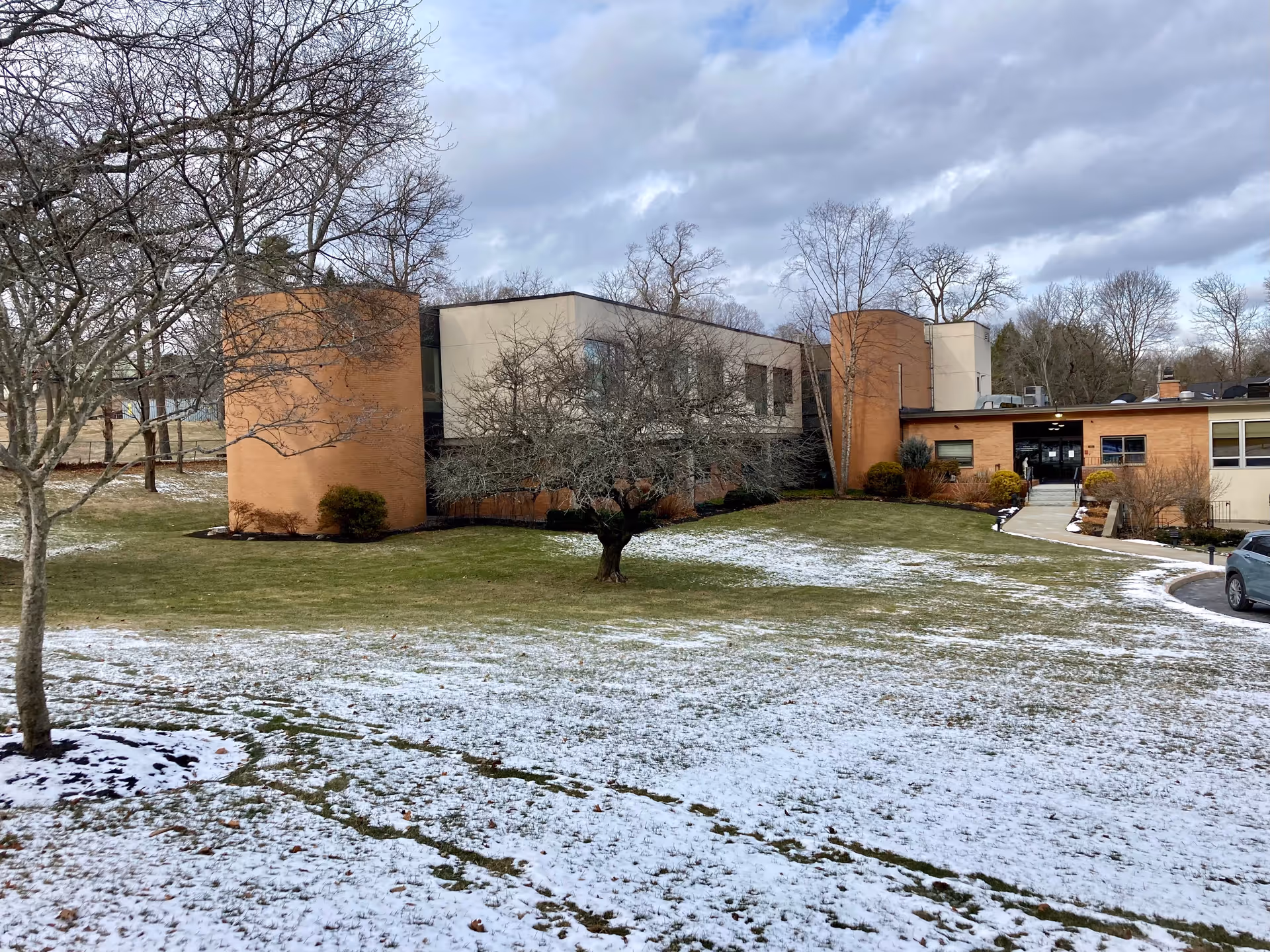 Low-rise nursing facility building with circular brick sections, a snow-dusted lawn, bare trees, and a driveway leading to the entrance.