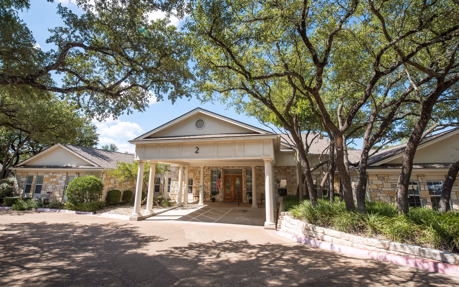 Exterior view of a single-story assisted living facility building with stone walls and a covered entrance supported by white columns. The building is surrounded by trees and landscaping under a clear blue sky.