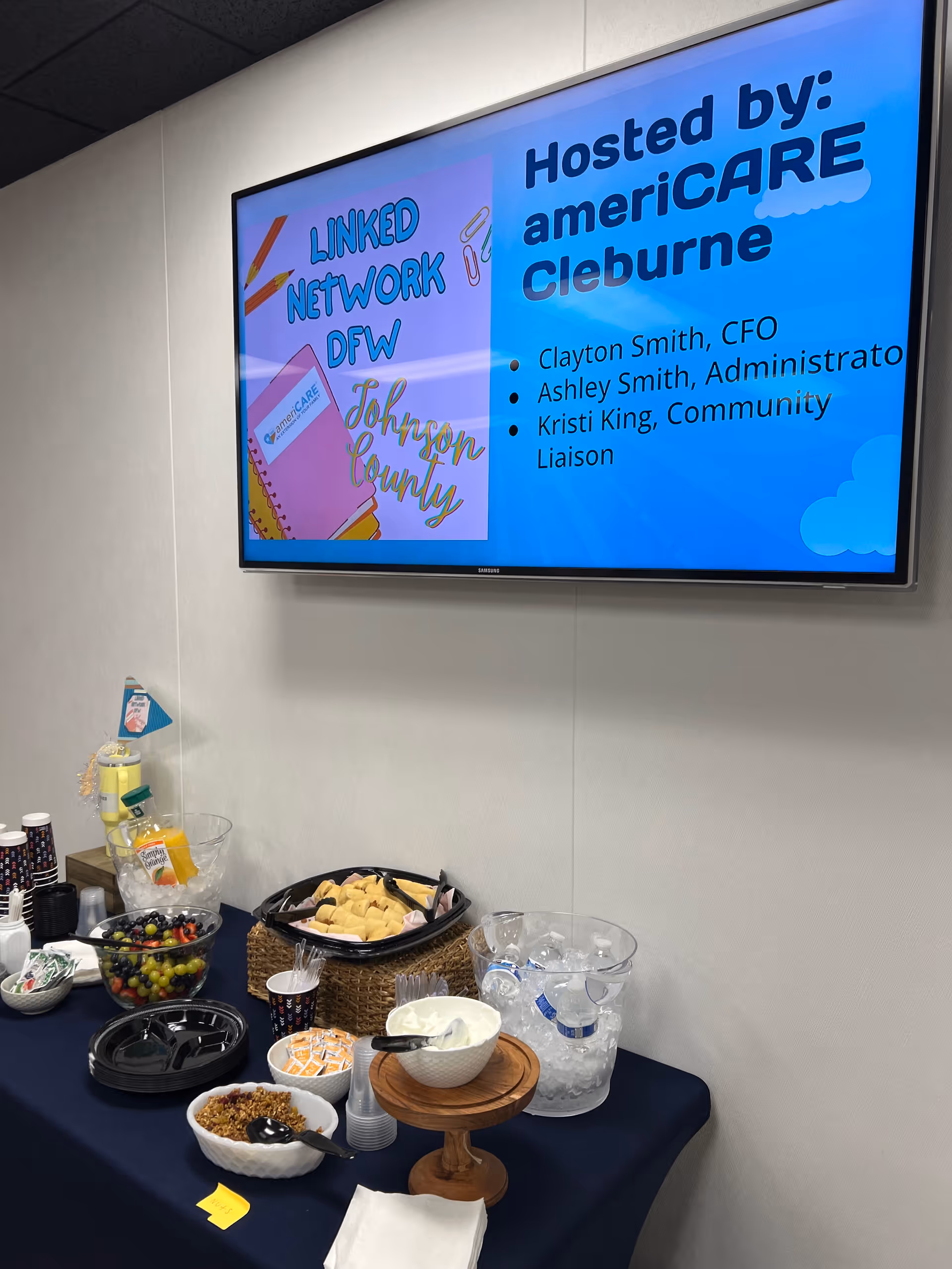 Indoor event setup with a snack table and a wall-mounted screen displaying ameriCARE Cleburne.