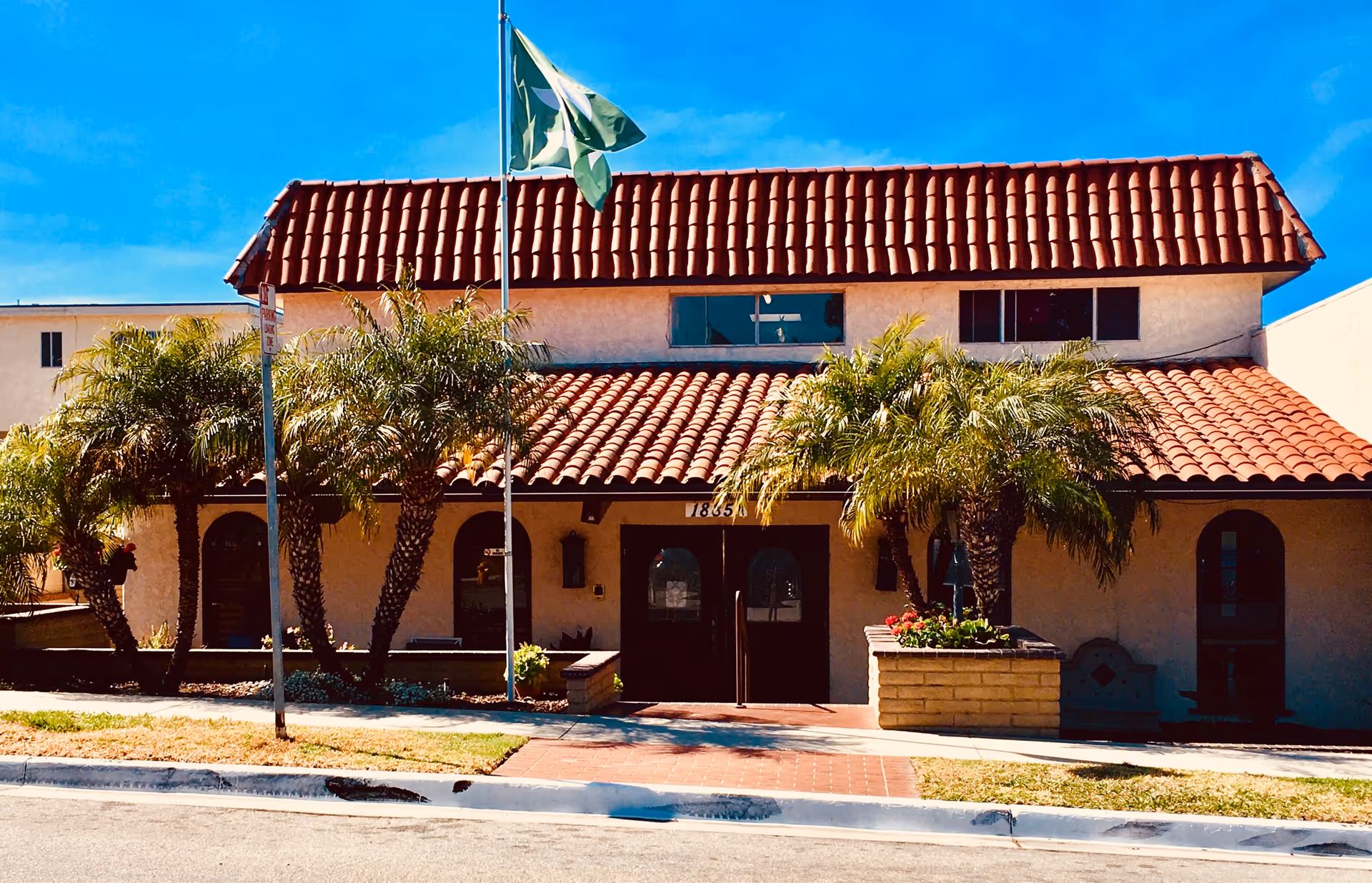 Exterior view of a Mediterranean-style assisted living building with palm trees and a flag.