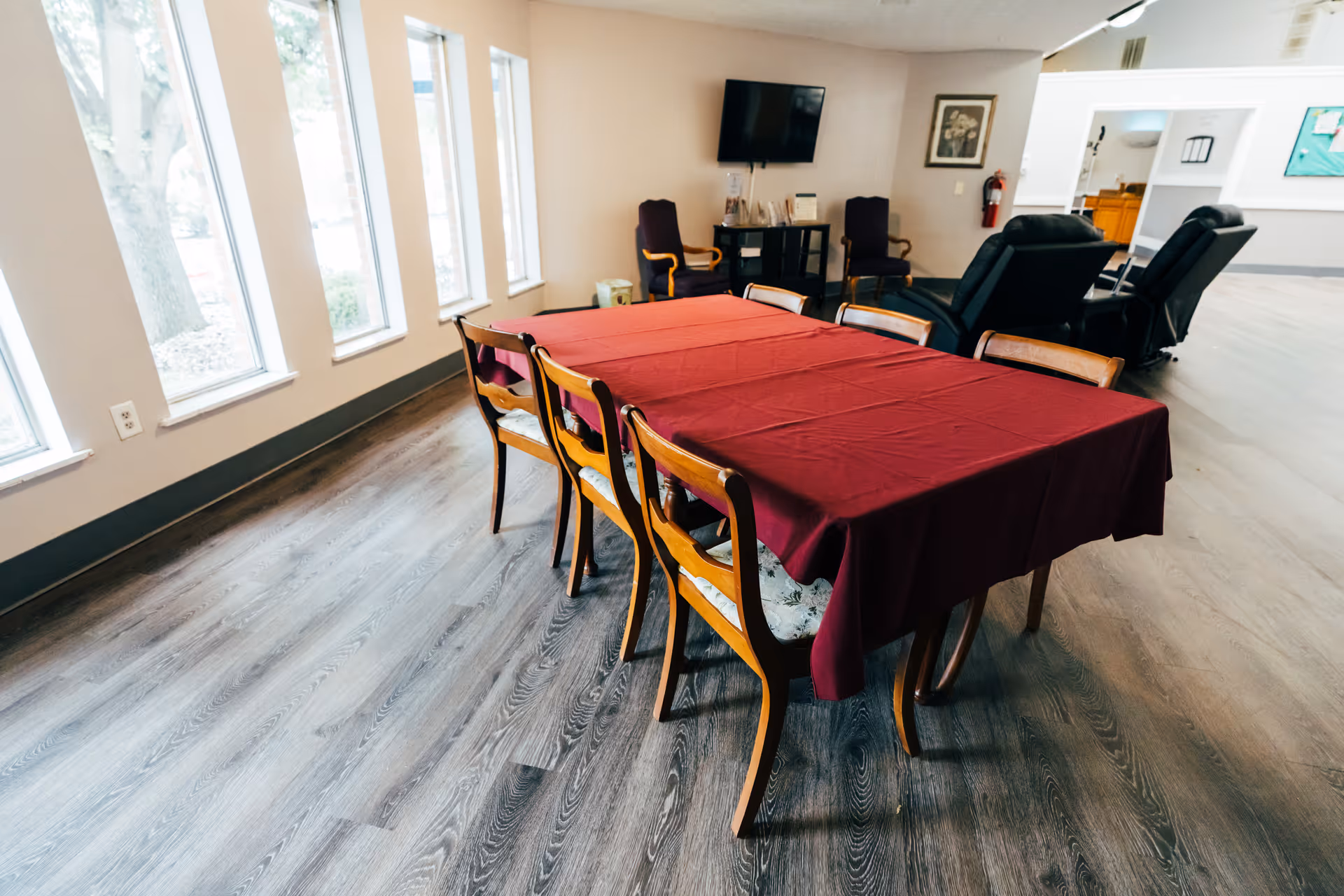 A communal dining table covered with a red tablecloth surrounded by wooden chairs in a bright common room with windows, reclining chairs, and a wall-mounted TV.