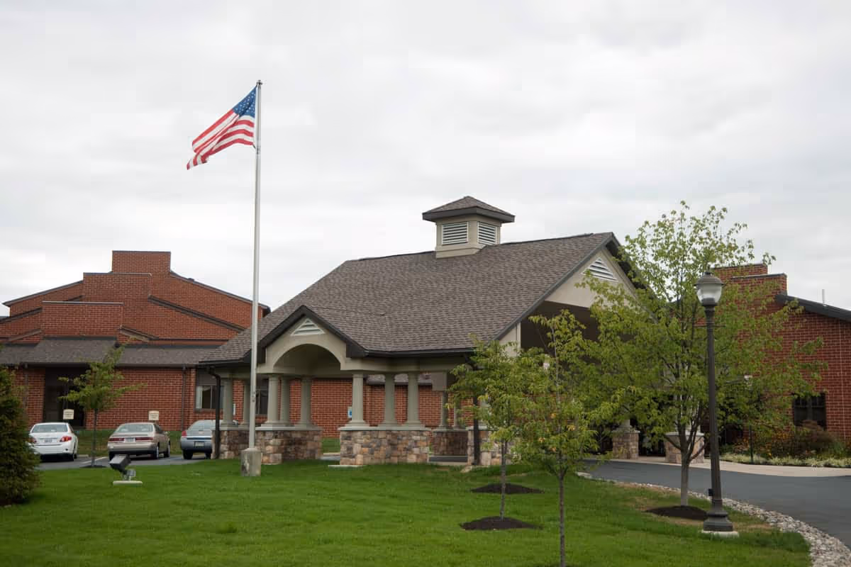 Brick senior living facility with flagpole, front entrance, cars, and landscaped lawn.