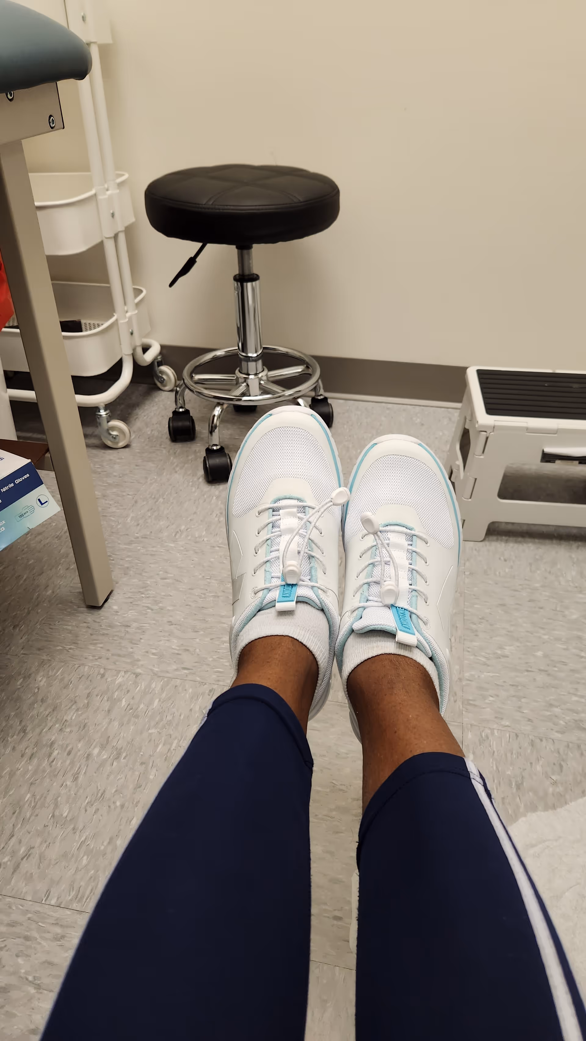 First-person view of sneakers in a medical exam room with stool and equipment nearby.