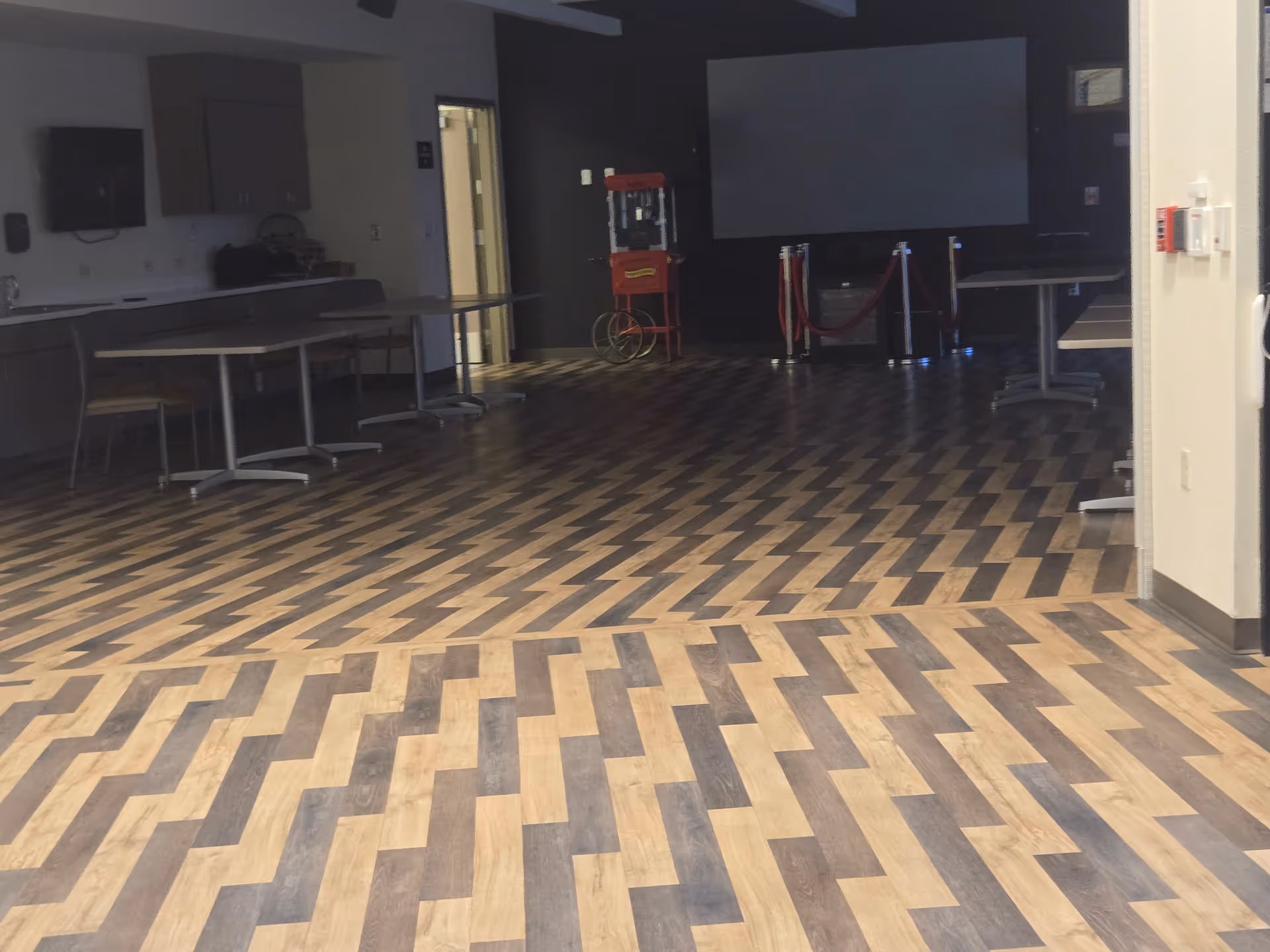 Empty multipurpose room with wood-pattern flooring, tables along the left, and a large screen.