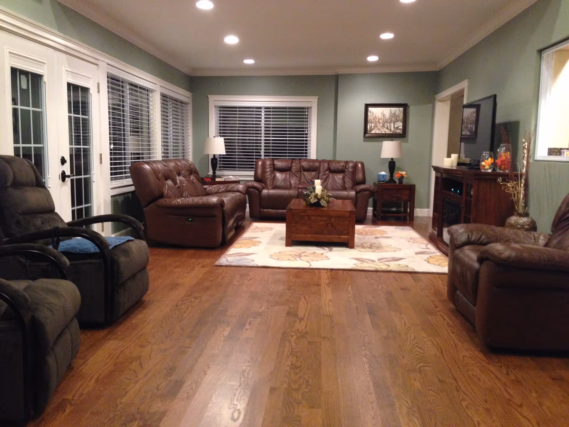 A cozy living room with wooden flooring, green walls, and recessed ceiling lights. The room features two brown leather sofas, two dark recliner chairs, a wooden coffee table on a floral area rug, two side tables with lamps, a TV on a wooden stand, and a window with white blinds.
