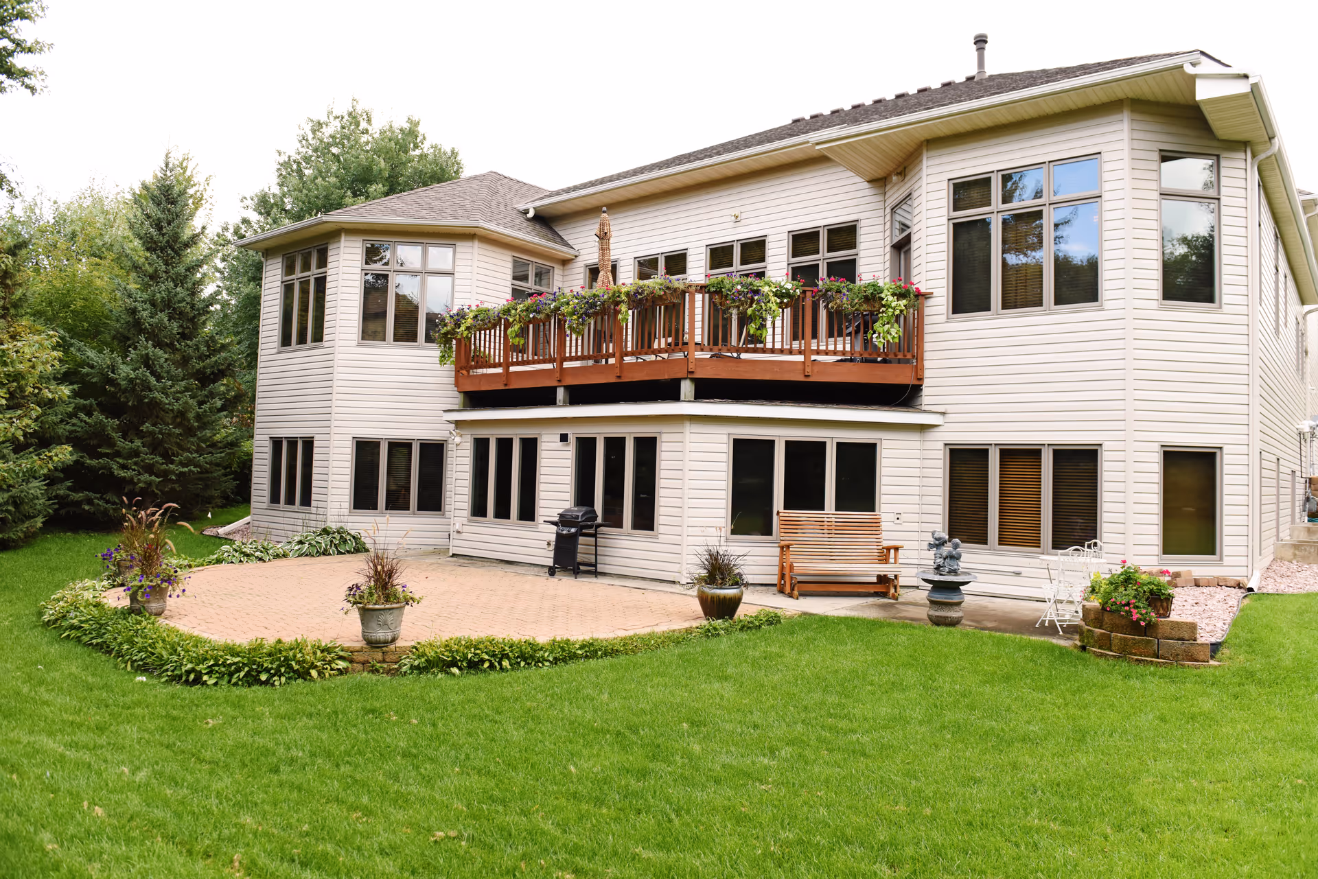 Two-story beige siding memory care building with a wooden balcony, ground-level patio, green lawn, and potted plants.