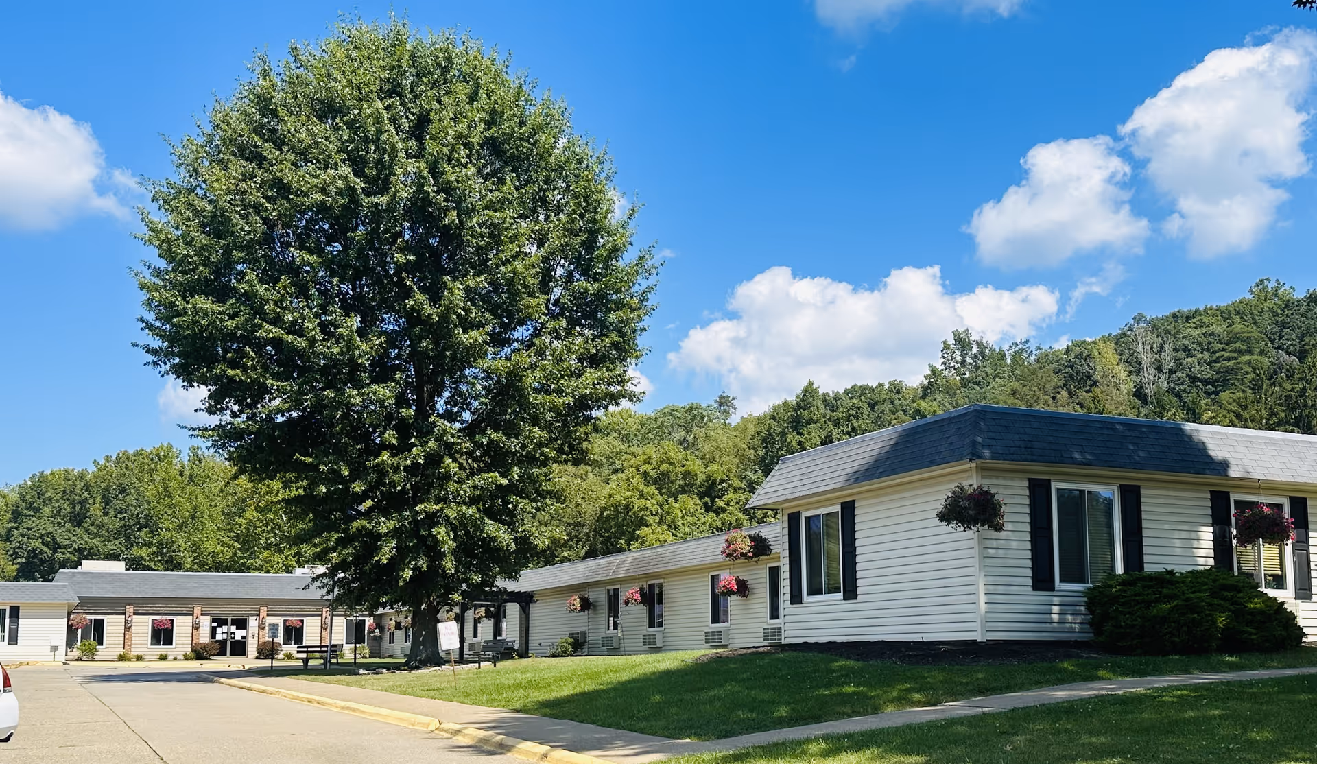 Exterior view of Worthington Healthcare Center on a sunny day with a large tree in the front lawn, white buildings with black shutters, hanging flower baskets, and a paved driveway.