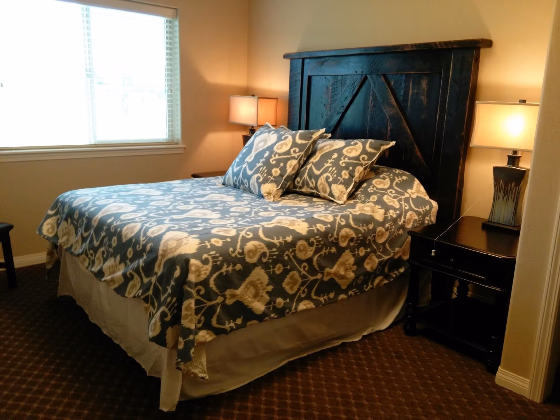A furnished bedroom with a patterned bedspread, dark wooden headboard, bedside tables and lamps beside a window.