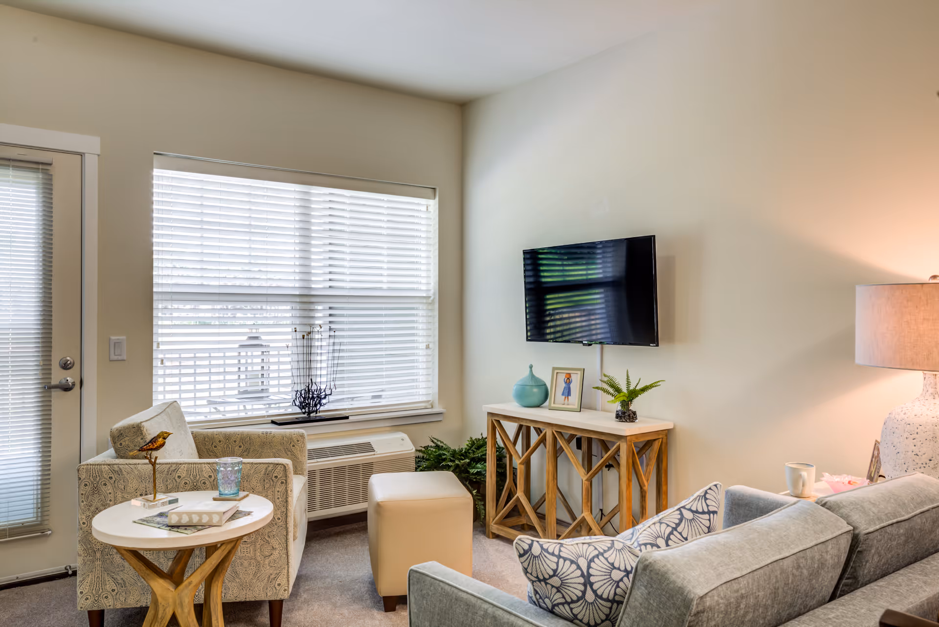 A cozy living room with a large window covered by white blinds, a patterned armchair next to a small round wooden table with a bird figurine and a glass, a beige ottoman, a gray sofa with a patterned pillow, a wooden console table with decorative items including a small plant and a framed photo, a wall-mounted flat-screen TV, and a large lamp on the right side.