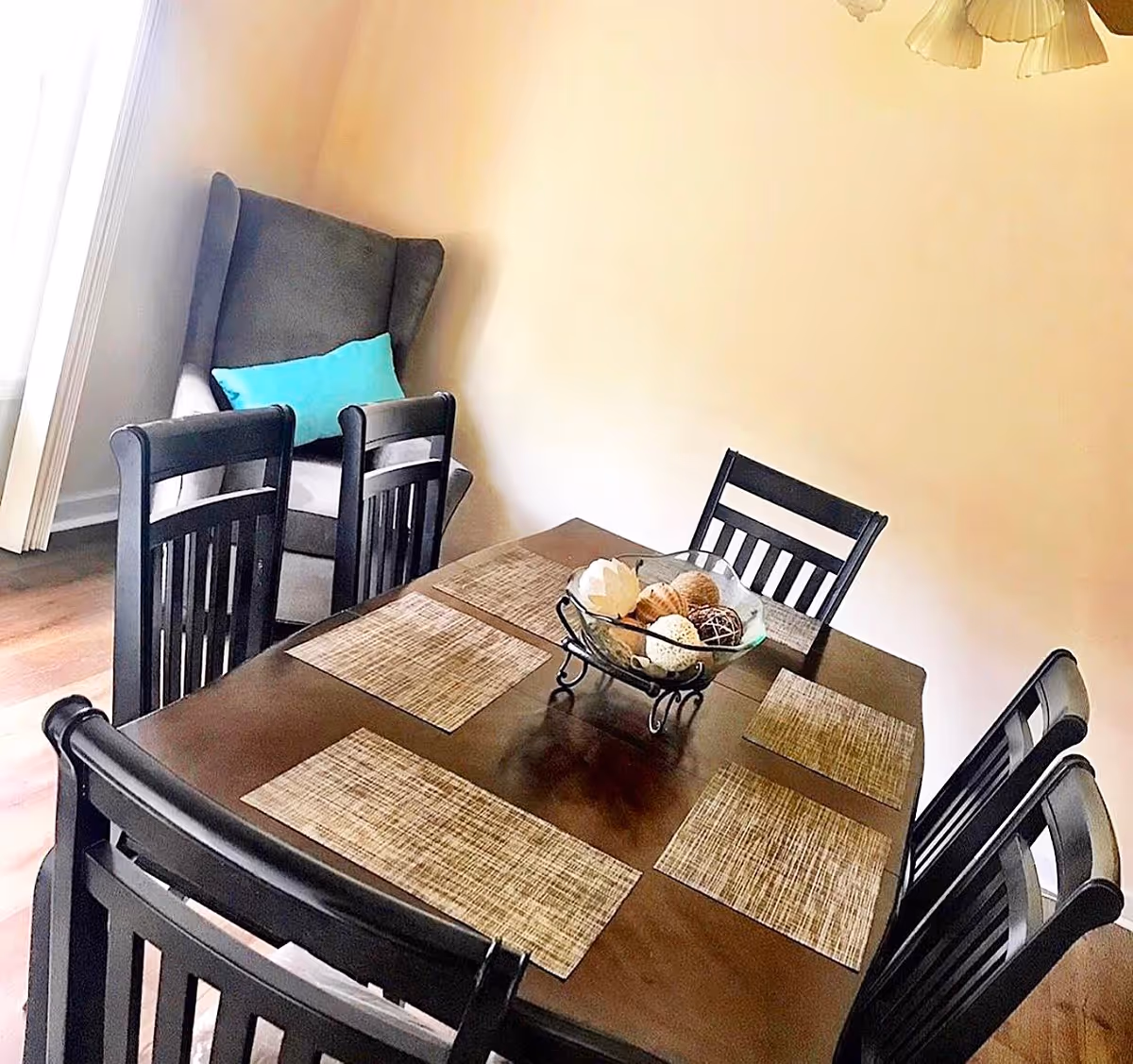 Wooden dining table with six black chairs, placemats and a decorative bowl, and a wingback chair with a turquoise pillow in the corner.