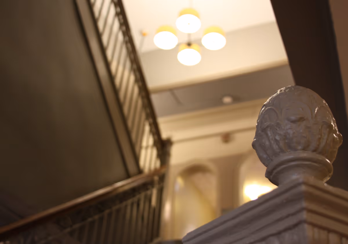 Close-up of a decorative stair banister finial and railing looking up toward a lit ceiling and upper landing.