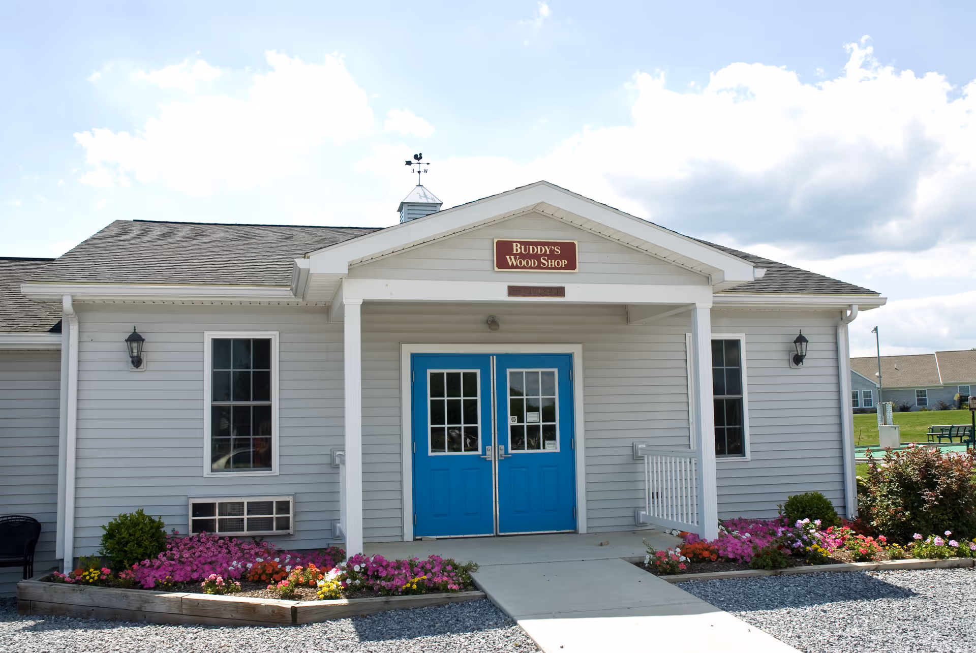 Exterior view of a single-story building with light gray siding and a dark shingled roof. The building has a small covered entrance with two bright blue doors and a sign above that reads 'Buddy's Wood Shop'. There are flower beds with colorful flowers on either side of the entrance and a gravel area in front. The sky is partly cloudy.