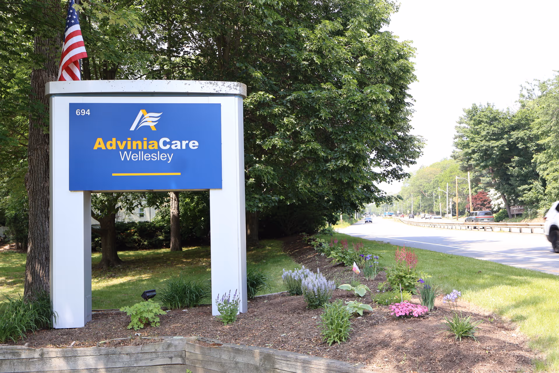 Outdoor view of a blue and white sign for AdviniaCare Wellesley, located on a grassy area with flowers and trees nearby, next to a road with cars driving by.