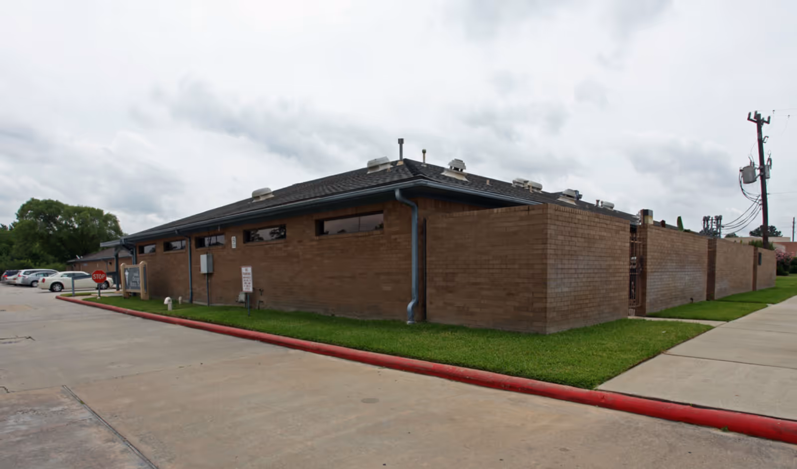 Exterior view of a single-story brick facility with grass, parking lot, and sidewalk under cloudy sky.