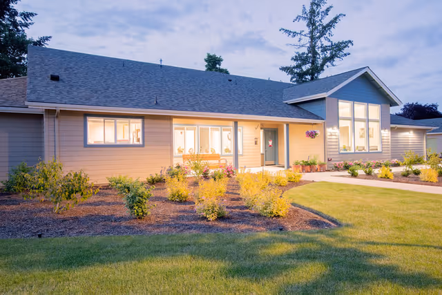 Exterior view of a single-story residential building with beige siding and a dark shingled roof, surrounded by a well-maintained lawn and landscaped garden beds with small shrubs and plants. The building has several large windows and a front door with a small porch area.