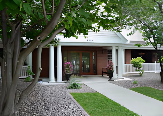 Front entrance of a single-story building with white columns, glass double doors, a concrete walkway and landscaped trees and shrubs.