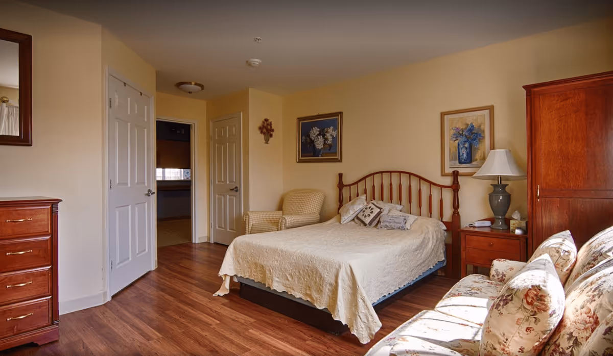 A cozy bedroom with a wooden bed frame, cream-colored bedspread, and decorative pillows. There is a floral armchair next to the bed, a wooden dresser with drawers on the left, and a wooden armoire on the right. The room has hardwood flooring, beige walls, two framed floral paintings, and a table lamp on a bedside table. A floral-patterned sofa is partially visible in the foreground.