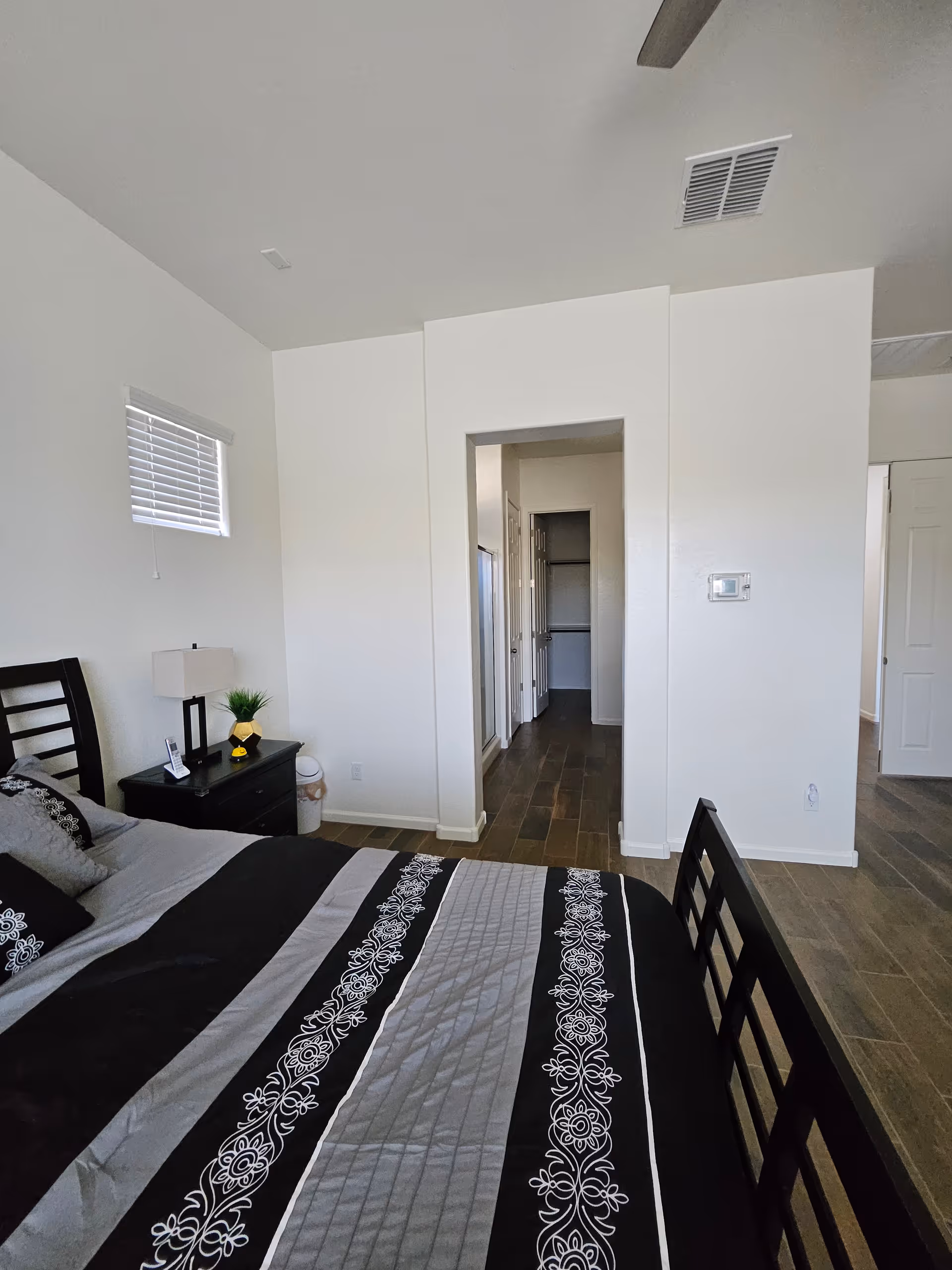 A bedroom in a senior living facility featuring a bed, nightstand, lamp, and hallway doorway.