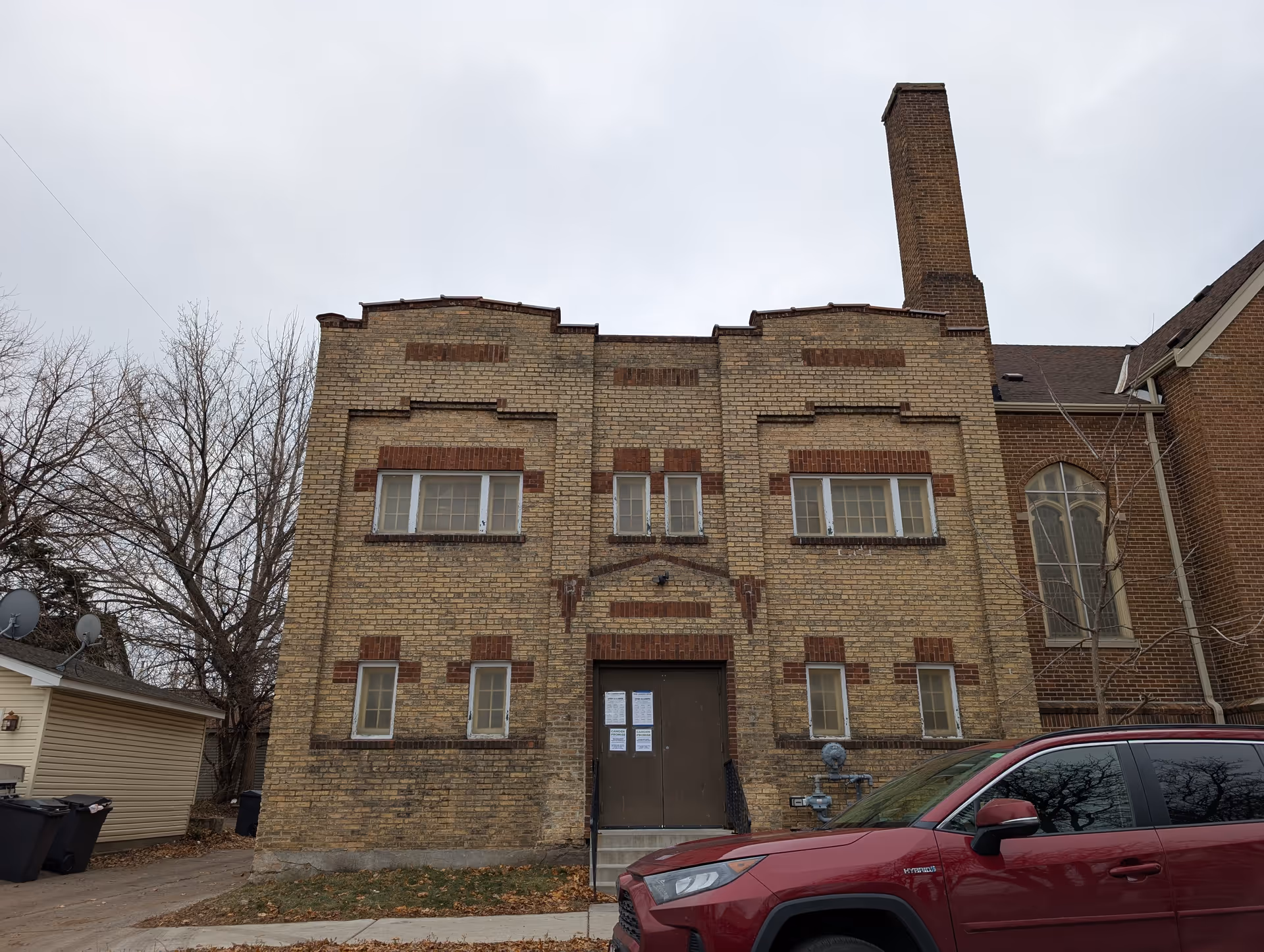 Front facade of a two-story yellow brick building with a tall chimney, small windows, and a red SUV parked in front.