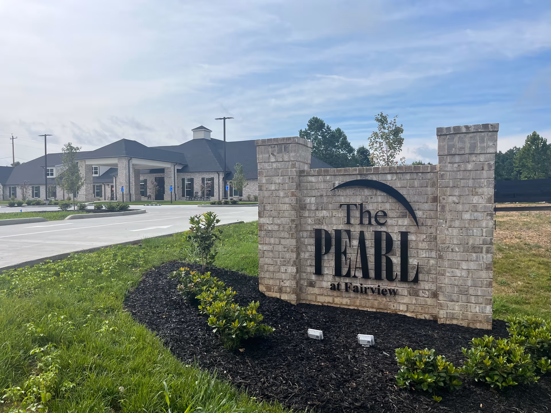 Exterior view of The Pearl at Fairview senior living facility showing a large brick sign with the facility name in front of a driveway and a single-story building with a covered entrance, surrounded by greenery and trees under a partly cloudy sky.