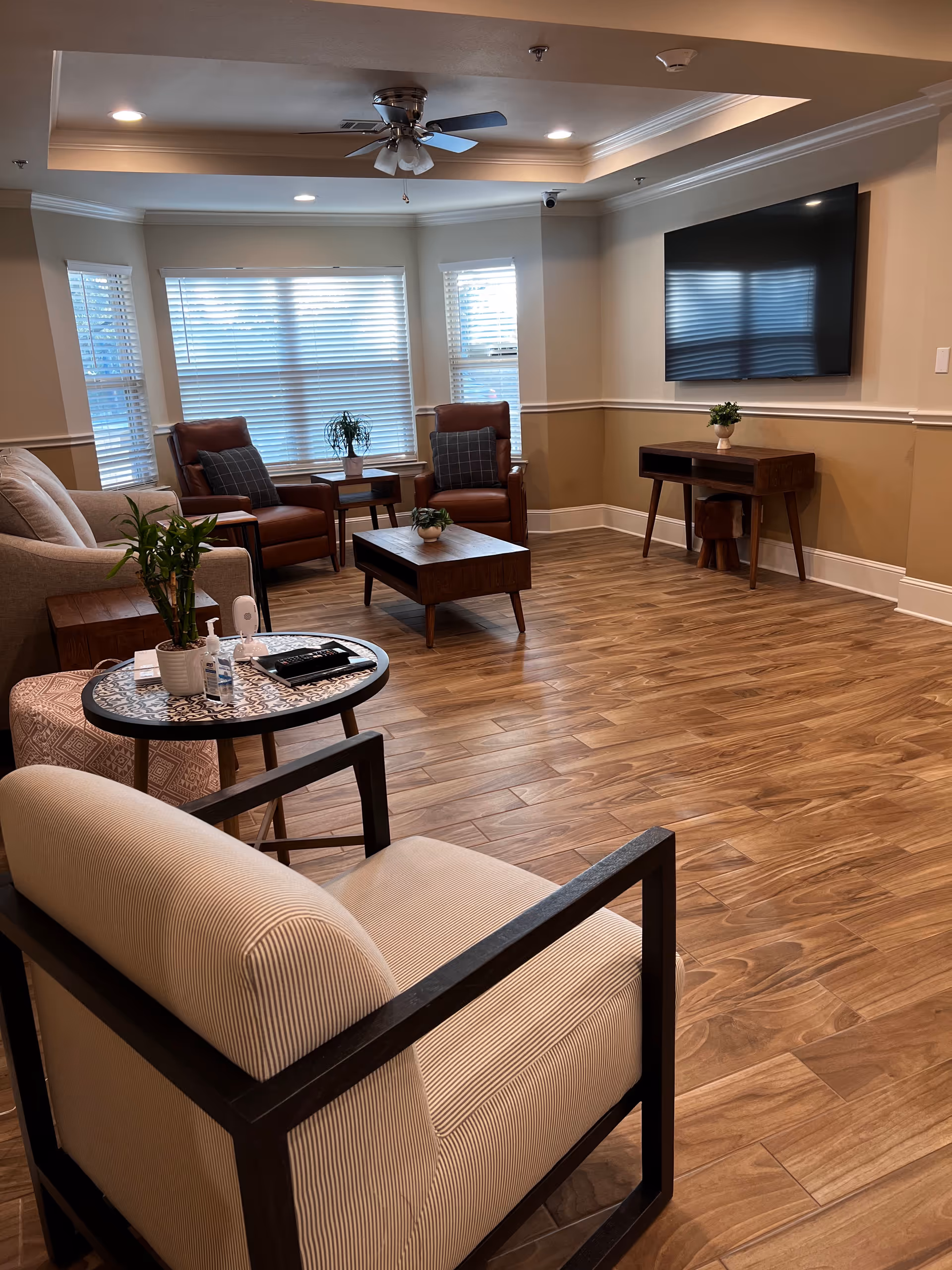 Cozy senior living lounge with beige walls, wood floors, and a wall-mounted TV.