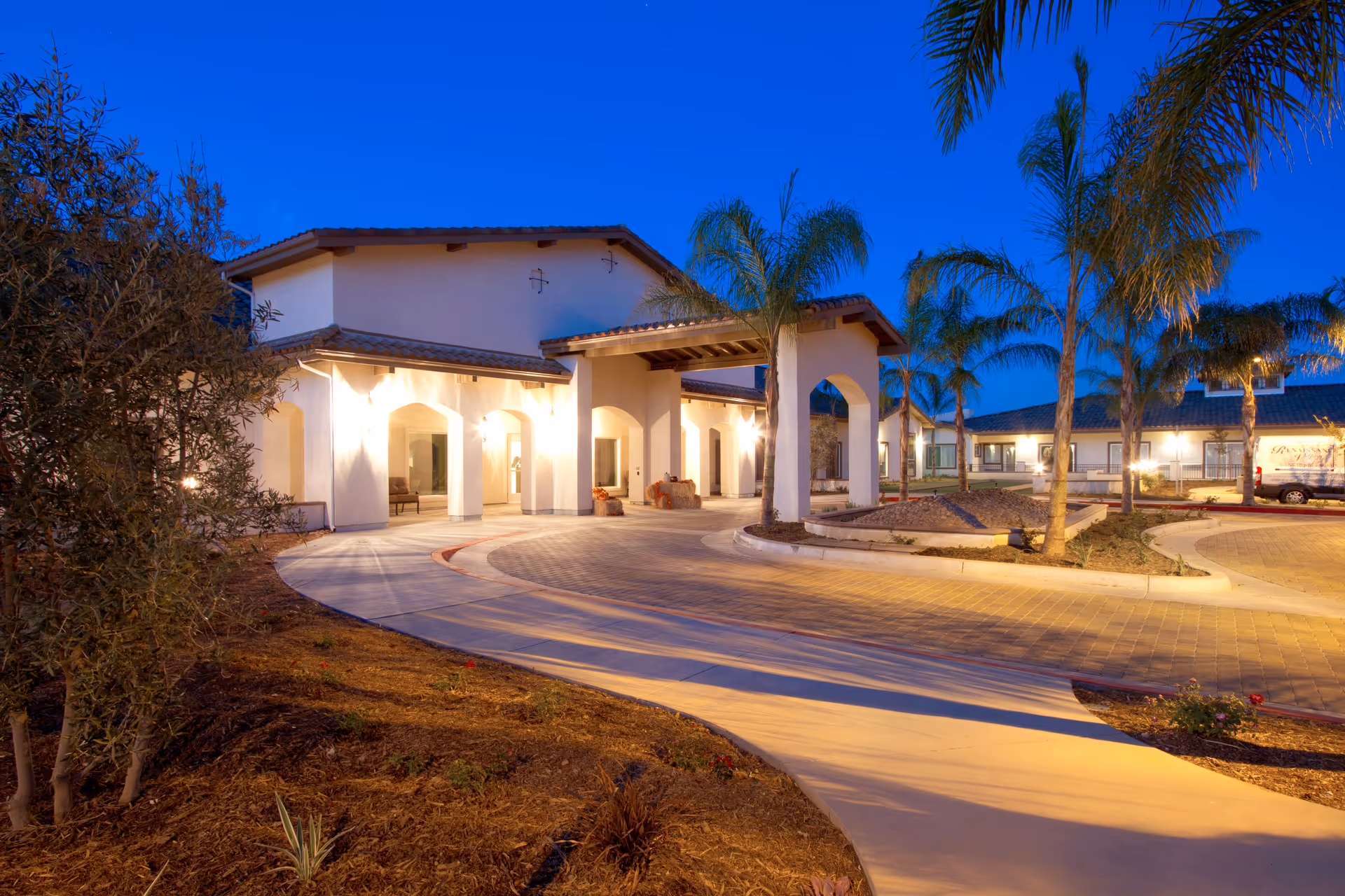 Exterior view of Manzanita Village senior living facility at dusk, showing a well-lit entrance with arches, palm trees, and a circular driveway.