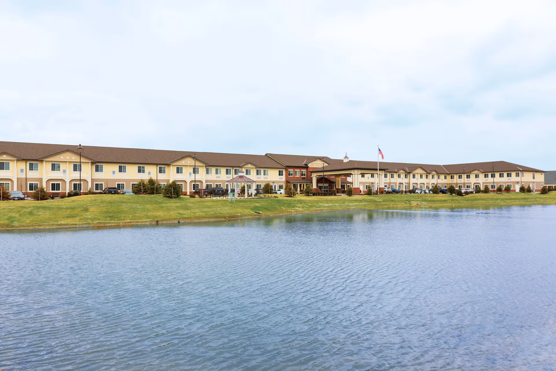 Wide exterior view of a large two-story senior living facility building with a brown roof and beige walls, situated behind a grassy area and a pond in the foreground under a cloudy sky.
