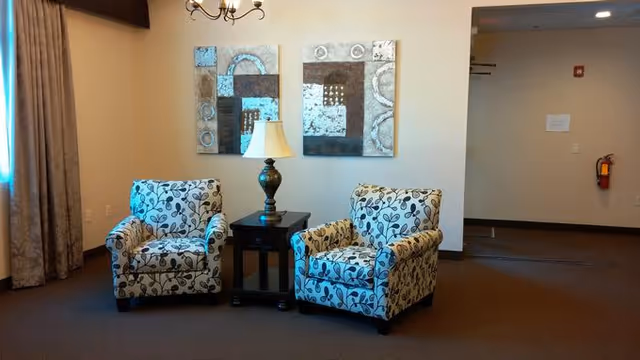 Two patterned armchairs with a floral design are placed on either side of a small dark wooden table with a lamp on it. Behind the chairs, there are two abstract paintings hanging on a beige wall. The room has brown carpet and beige walls with a window covered by curtains on the left side.