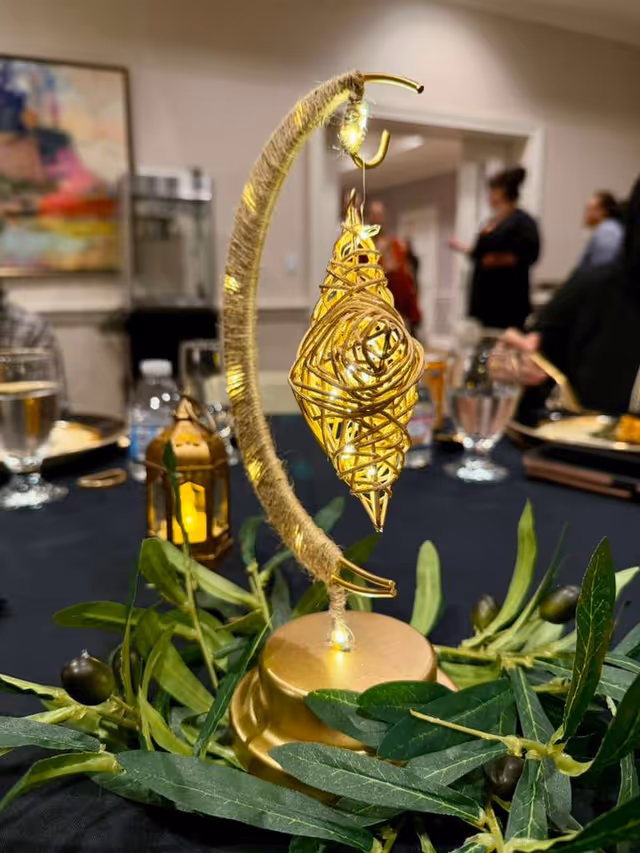 Close-up gold centerpiece on a dining table with olive garland, blurred guests behind.