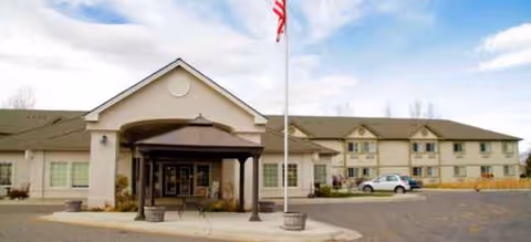Front view of a senior living facility with entrance canopy, flagpole, and parked cars.