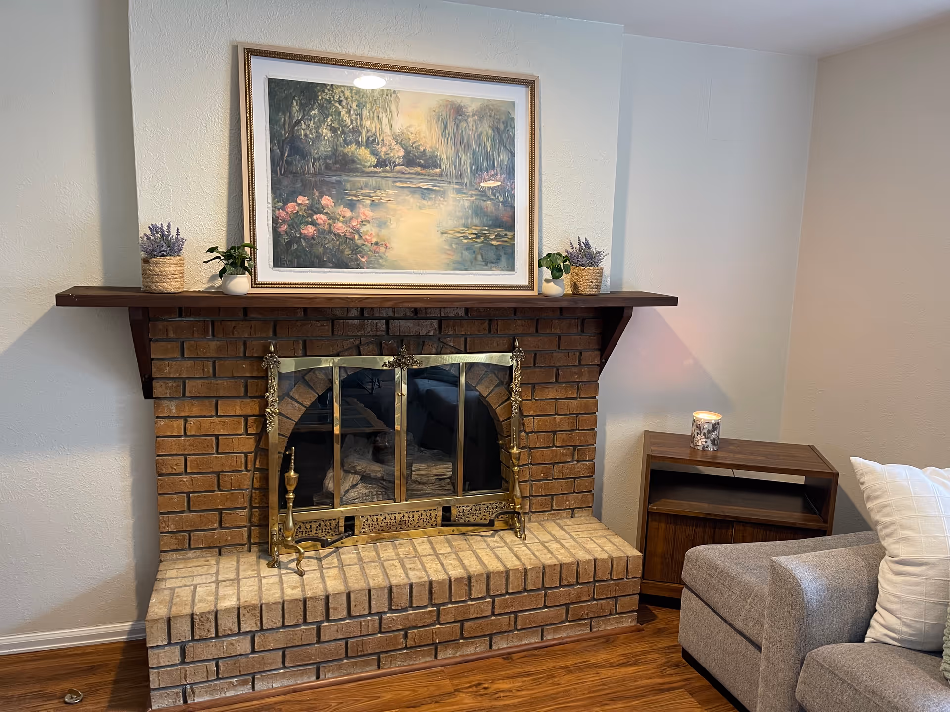 Brick fireplace with brass screen, mantel painting, plants, and a side table.