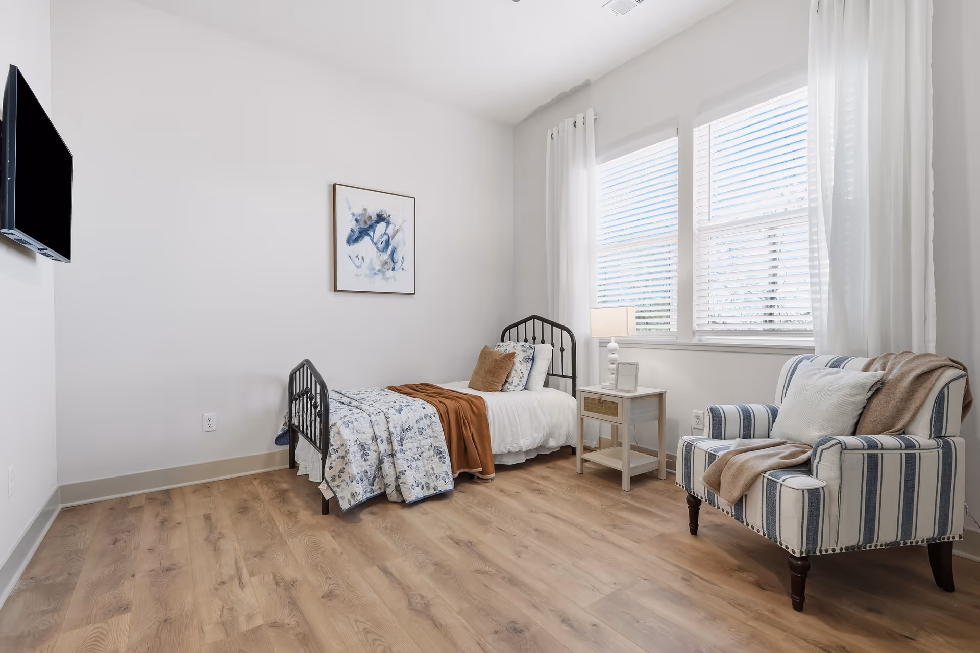 A bright bedroom with a single bed, nightstand, striped chair, and large windows.